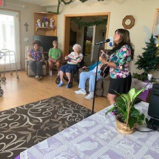 A person is playing guitar for a group of seniors in a nursing home living room.