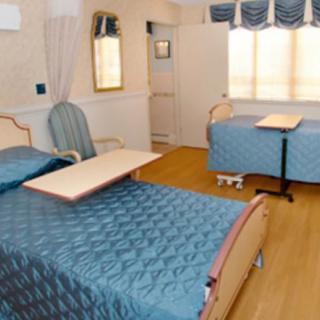 A hospital room with two patient beds, equipped with adjustable tables and a comfortable chair.