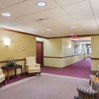 A well-lit hallway with chairs and a table against the wall, featuring carpet flooring.