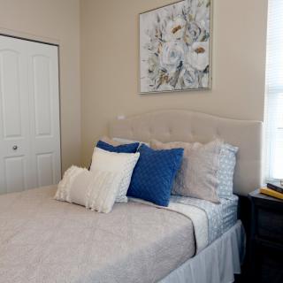A cozy bedroom featuring a neatly made bed with white and blue pillows, adorned by a floral painting above and natural light streaming through the window.