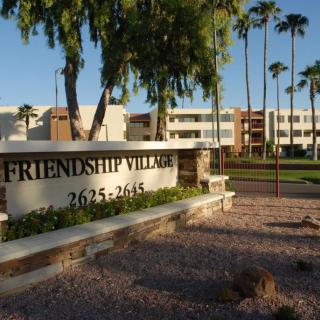 A view of Friendship Village sign with palm trees and residential buildings in the background.