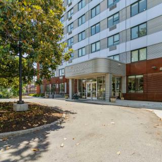 The image shows the entrance of a modern apartment building surrounded by trees.