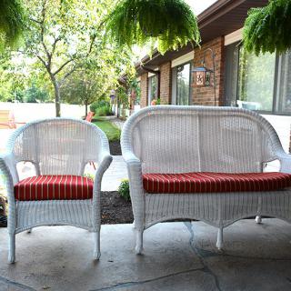 A wicker chair and loveseat with red cushions on a patio surrounded by greenery.