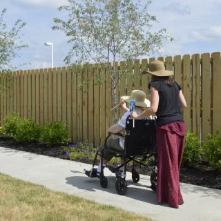 A caregiver assists an elderly person in a wheelchair along a garden path.