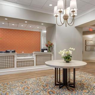 A welcoming hotel lobby with a central table, floral decorations, and a well-lit reception desk.