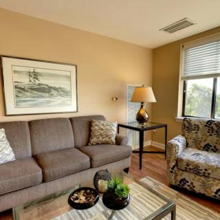 A cozy living room with a sofa, armchair, and coffee table, adorned with artwork and a lamp.