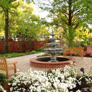 A serene garden with a central fountain surrounded by benches, trees, and blooming flowers.