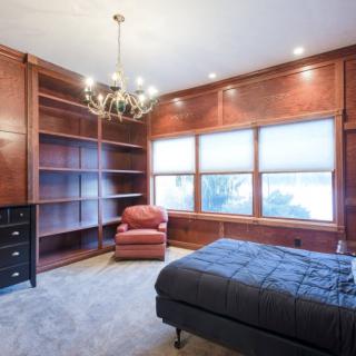 A cozy bedroom with wood paneling, featuring built-in bookshelves, a comfortable armchair, a bed, and a chandelier providing elegant lighting.