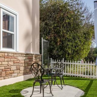 A cozy outdoor patio area with a small table and two chairs on artificial grass, surrounded by a white picket fence.