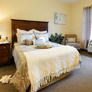 A cozy bedroom with a well-decorated bed, nightstand, and a comfortable chair by the window.