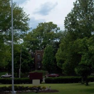 A peaceful park scene with a flagpole and memorial surrounded by lush green trees under a cloudy sky.