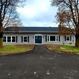 A single-story house with a central blue door, surrounded by trees and a driveway leading up to it.