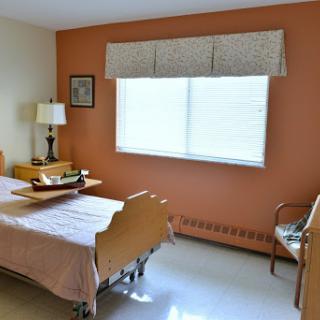 A cozy bedroom with a single bed, a lamp, and a chair, featuring warm wall colors and natural light from the window.