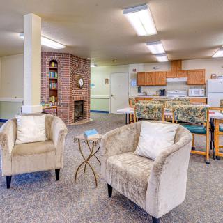 A cozy living room with plush chairs, a small table, and an open kitchen area in the background.