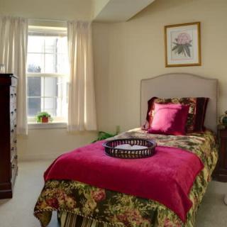A cozy bedroom with a single bed adorned with floral bedding and a vibrant pink blanket, illuminated by sunlight through the window.