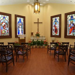 A small chapel interior featuring stained glass windows, an altar with a cross, and arranged chairs.