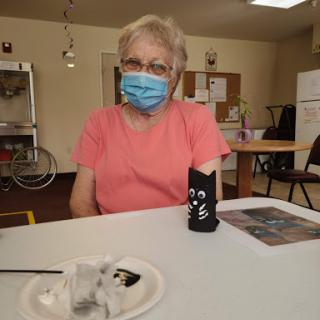 A senior woman wearing a mask sits at a table, engaged in a social activity.
