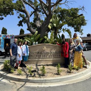 A group of people stands around a sign in a sunny outdoor setting with a large tree in the background.