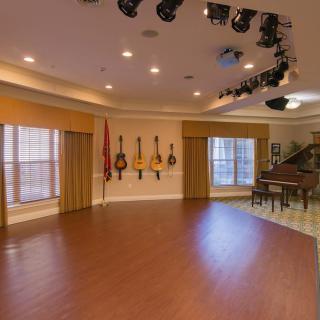 A spacious music room featuring a selection of guitars on the wall, a piano, and overhead stage lighting.