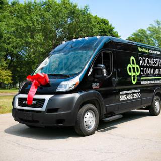 A black delivery van adorned with a large red ribbon, parked outdoors surrounded by greenery.