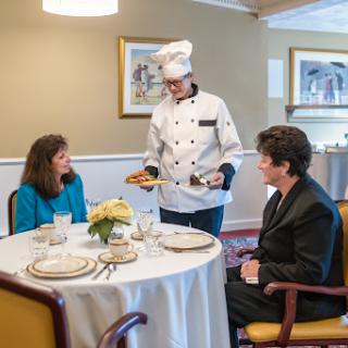 A chef serving a meal to two guests at an elegantly set dining table.