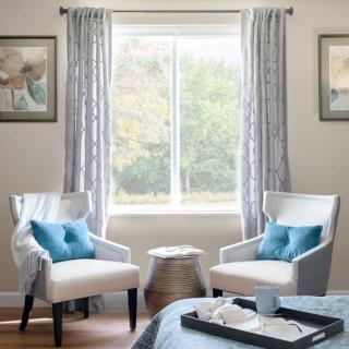 A cozy living room corner featuring two white chairs with blue pillows, a small table, and natural light streaming in through the window.