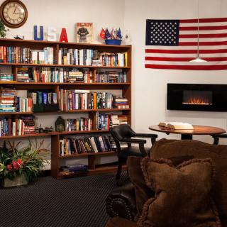 A cozy reading room featuring a large bookshelf filled with books, an American flag on the wall, and a comfortable armchair near a fireplace.