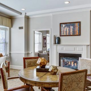 A cozy living room with a round dining table, chairs, and a fireplace, adorned with artwork and large windows.