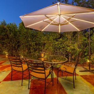 An inviting outdoor patio area with a lit umbrella and a cozy seating arrangement, surrounded by lush greenery at night.