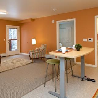A cozy living room with orange walls, featuring a small table and stools, a lamp, and a doorway to the outside.