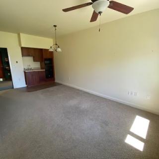 A spacious, empty living room featuring a ceiling fan and access to the kitchen area.