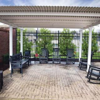 A tranquil outdoor patio with a pergola, featuring rocking chairs and benches on a brick floor.