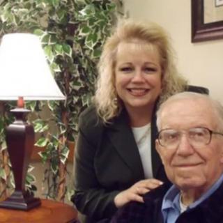A woman smiles while standing next to an elderly man in a cozy room.