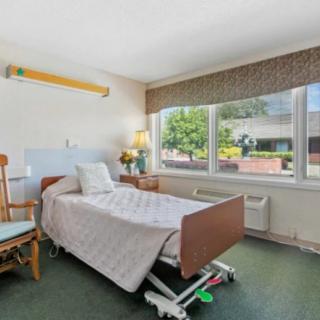 A well-lit hospital room with a single bed, chair, and a large window offering an outside view.