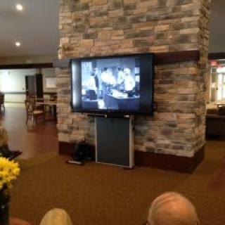 A group of people watching a black-and-white movie on a television mounted on a brick wall in a cozy indoor setting.