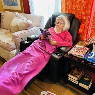An elderly woman is comfortably reading in a recliner, wrapped in a pink blanket in her cozy living room.