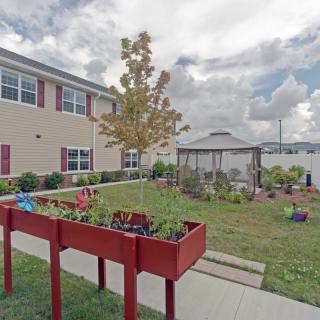 A well-maintained garden area with raised beds and a gazebo near a building under a partly cloudy sky.