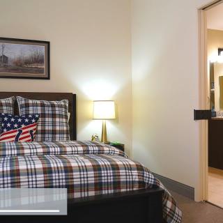A cozy bedroom with a plaid bedding set, an American flag cushion on the bed, and a lamp on a side table. The room also features wall art and an open doorway.