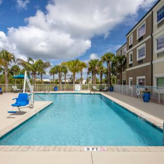 An outdoor swimming pool surrounded by a fence with palm trees and a multi-story building in the background under a cloudy sky.