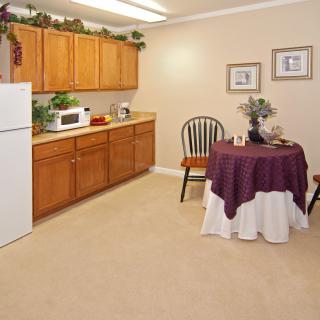 A cozy kitchen and dining area with wooden cabinets, a refrigerator, microwave, and a small round table draped with a purple and white tablecloth.