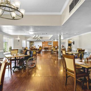 A spacious dining area with wooden flooring, equipped with tables and chairs, featuring a chandelier and a wheelchair for accessibility.