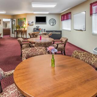A cozy community room with round wooden tables and patterned chairs, featuring decorative vases, a television, and a wall clock.