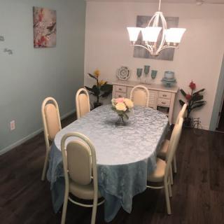 A cozy dining room with an oval table covered by a light blue tablecloth, surrounded by chairs and lit by a chandelier.