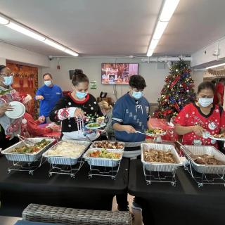 People serving food at a festive Christmas buffet, wearing holiday-themed clothing and masks.