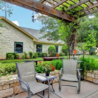 A tranquil patio space with chairs under a pergola, surrounded by lush garden greenery beside a stone house.