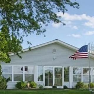 A white house with an American flag flying in front, surrounded by a neatly trimmed lawn and trees under a partly cloudy sky.