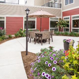 A cozy outdoor patio area with a table, chairs, and an umbrella surrounded by vibrant flowers and landscaping.
