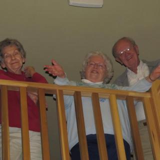 Three seniors smiling and waving from a balcony indoors.