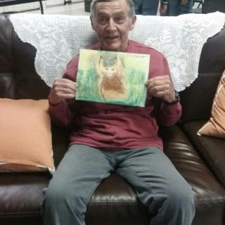 An elderly man sitting on a sofa, holding up a painting.