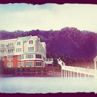 A scenic view of a house by the lake with a pier extending over the water, set against a backdrop of lush forested hills.